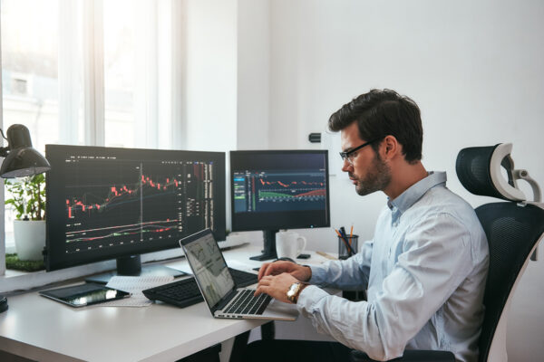 Workplace of trader. Young bearded trader wearing eyeglasses using his laptop while sitting in office in front of computer screens with trading charts and financial data. Stock exchange. Financial trading concept. Investment concept